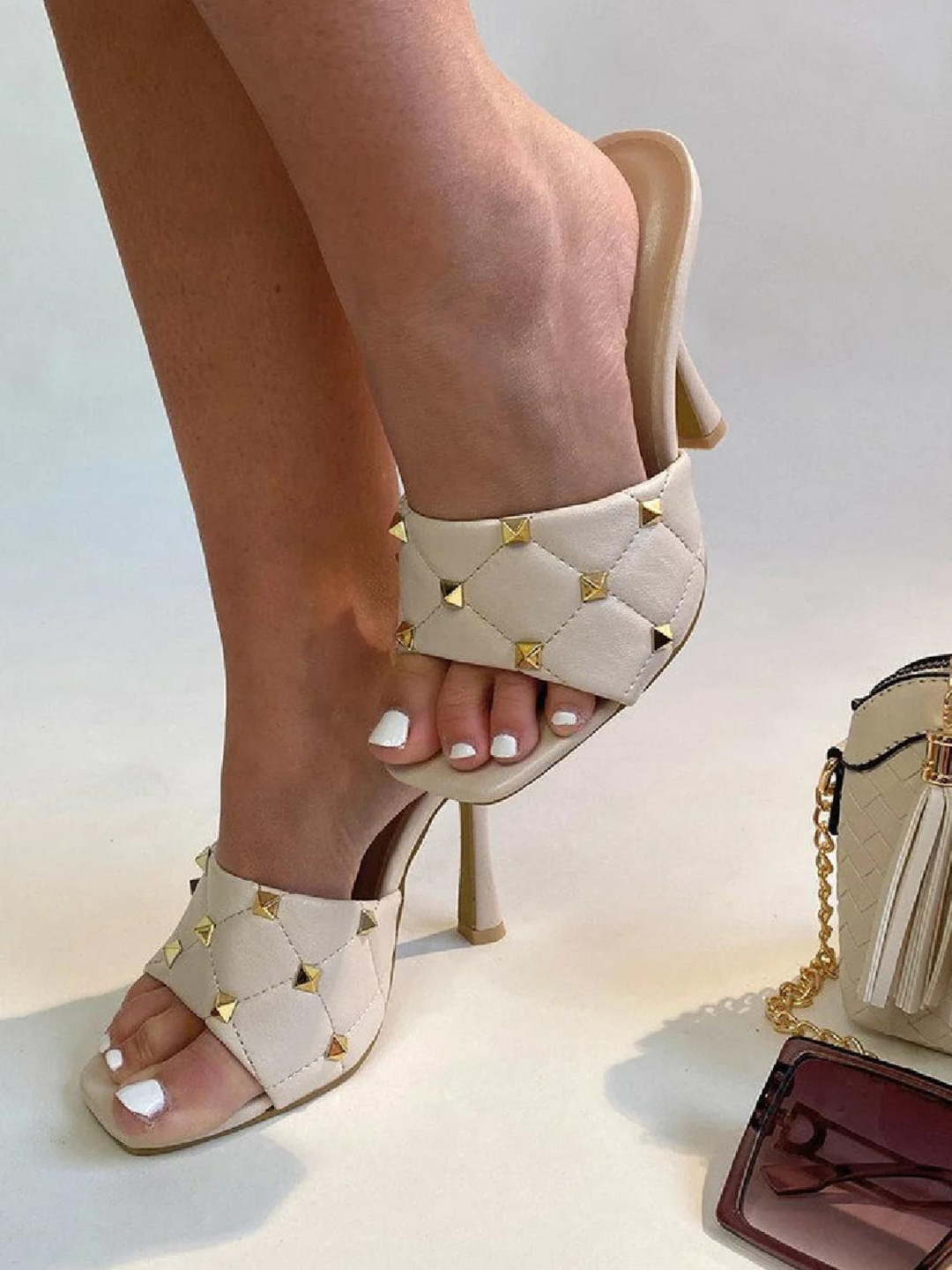 Cream quilted faux leather mule heels with gold studded heels. Model waers cream studded mule heels, she lifts obe leg above the ground. A hand bag and sunglasses are on the ground.