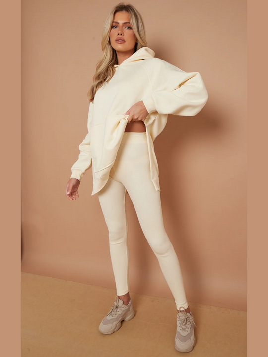 Full length shot. Model wears oversized cream hoodie with matching ribbed leggings. Model has blonde hair, she  lifts the jumper and shows her ribbed leggings. Model wears chunky trainers.