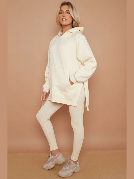 Full length. Model wears oversized cream hoodie with matching ribbed leggings. Model stands to the side, she places one hand in her pocket and the other by her side. The oversized jumper and ribbed leggings can be seen. The model wears a chunky trainers.