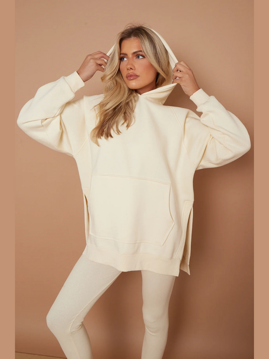 Model wears oversized cream hoodie with matching ribbed leggings.  Model wears her hood on her head and touches her hoodie on either side of her head. Model looks off into the distance.