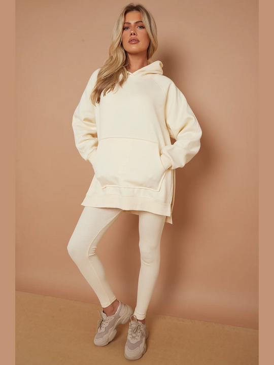 Full length shot. Model wears oversized cream hoodie with matching ribbed leggings.  Model has blonde hair, she stands with both hands in her jumper and bends one knee slightly. Model looks directly into the camera, she wears chunky cream trainers.