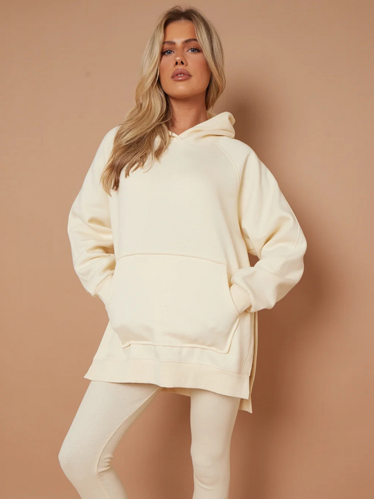 Model wears oversized cream hoodie with matching ribbed leggings. The model has blonde hair, she faces the camera and places one leg to the side. The model places both hands in her pockets and looks directly into the camera.