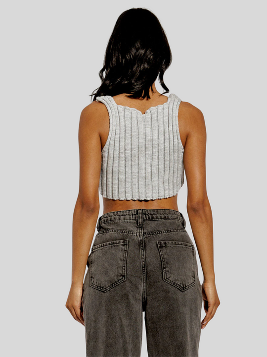 The model wears  a stylish grey knitted crop top with an asymmetric hem. The crop top, made from 100% acrylic, boasts a modern and trendy design with its uneven hemline adding a touch of asymmetry. Model has light brown skin and black hair, she stands with her back to the camera and her hands by her sides. The back of the knited crop top can be seen, as well as the back of the dark grey jeans.