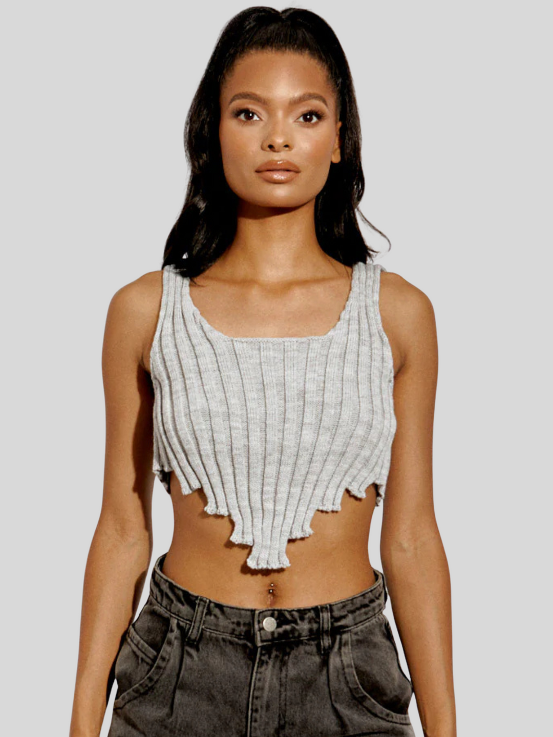 Model wears a stylish grey knitted crop top with an asymmetric hem. The crop top, made from 100% acrylic, boasts a modern and trendy design with its uneven hemline adding a touch of asymmetry. Model has light brown skin and black hair.