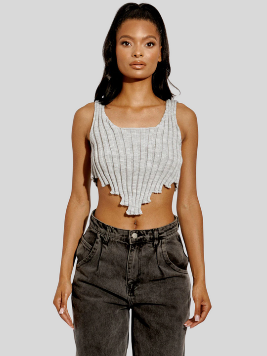 This image features a stylish grey knitted crop top with an asymmetric hem. The crop top, made from 100% acrylic, boasts a modern and trendy design with its uneven hemline adding a touch of asymmetry. Model has light brown skin and black hair, she stands facing the camera, placing  both hhands by her side, the front of the top can be seen.