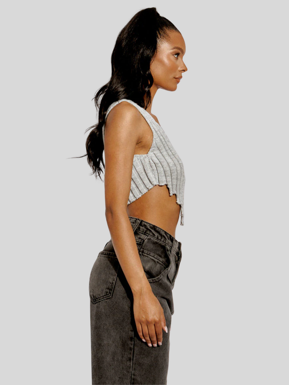 The model wears  a stylish grey knitted crop top with an asymmetric hem. The crop top, made from 100% acrylic, boasts a modern and trendy design with its uneven hemline adding a touch of asymmetry. Model has light brown skin and black hair, she stands to the side with her arm by her side. The side of the top can be seen , including the  sid of the asymmetric  hem.