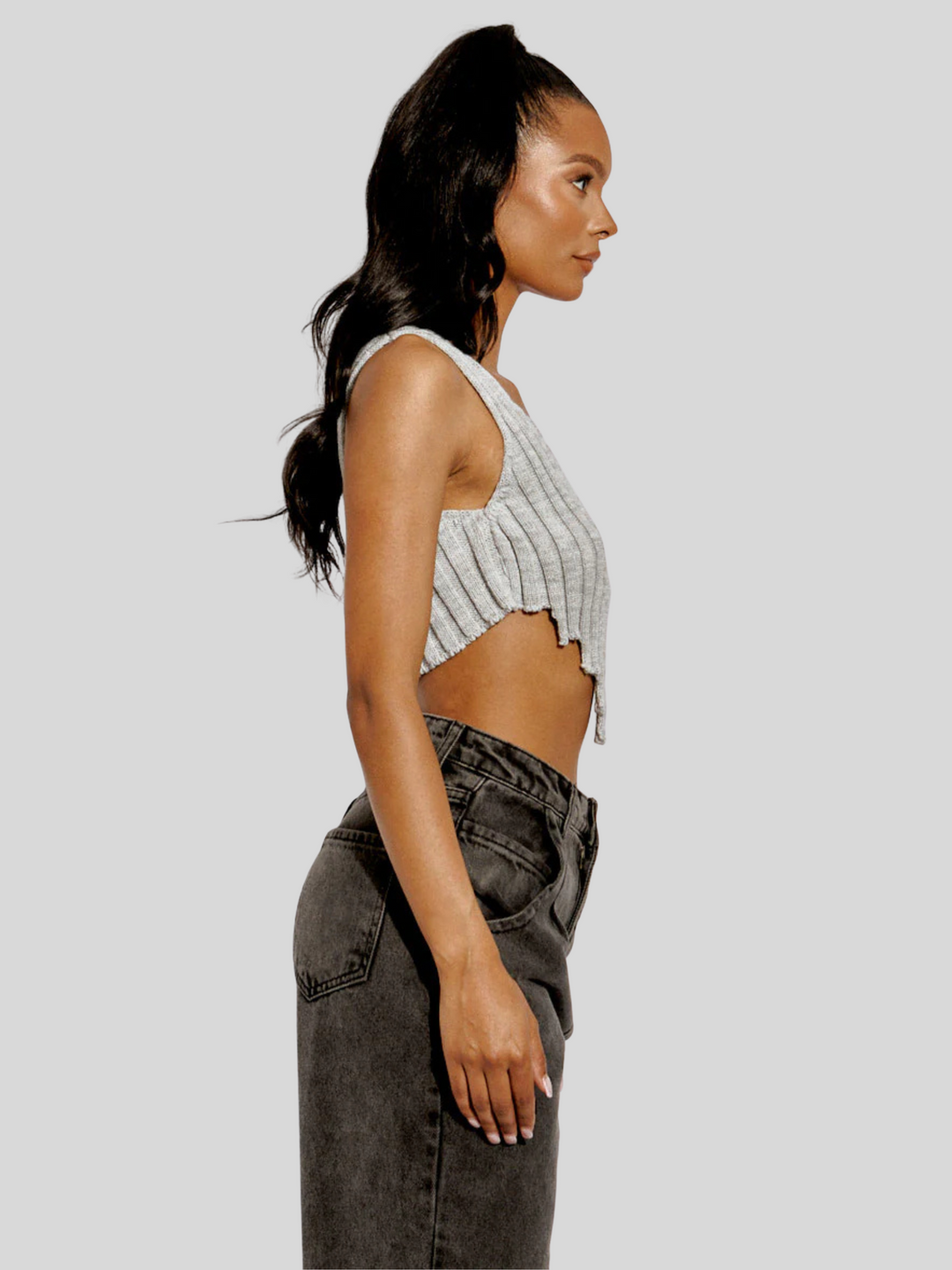 The model wears  a stylish grey knitted crop top with an asymmetric hem. The crop top, made from 100% acrylic, boasts a modern and trendy design with its uneven hemline adding a touch of asymmetry. Model has light brown skin and black hair, she stands to the side with her arm by her side. The side of the top can be seen , including the  sid of the asymmetric  hem.