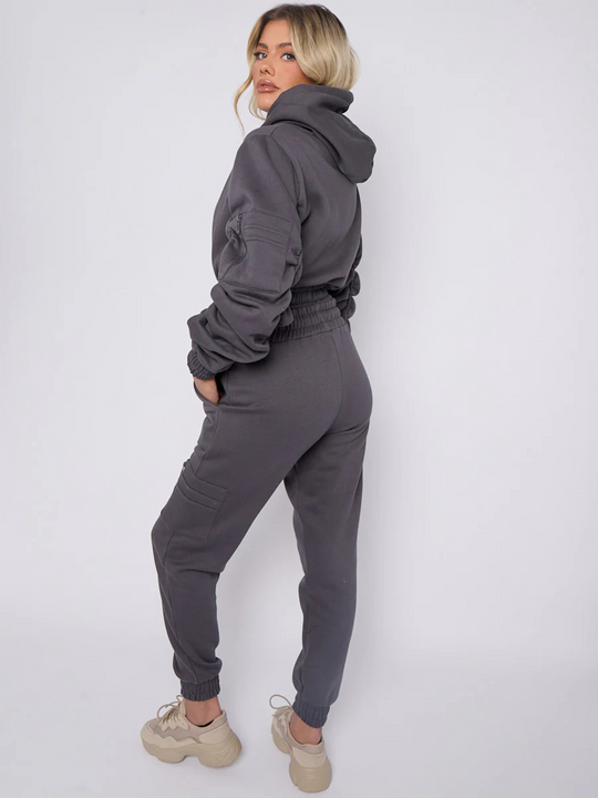 Full length shot. Model wears grey hoodie and matching fitted joggers set. The hoodie has a draw string design, front zip fastening, elasticated waist and ruched sleeves. The matching joggers too has an elasticated waist. The model has blonde hair, she stands to the side and places  her hand in her pocket. Mdel turns her head so that she is looking into the camera, he wears chunky trainers that compliments her jogger set.