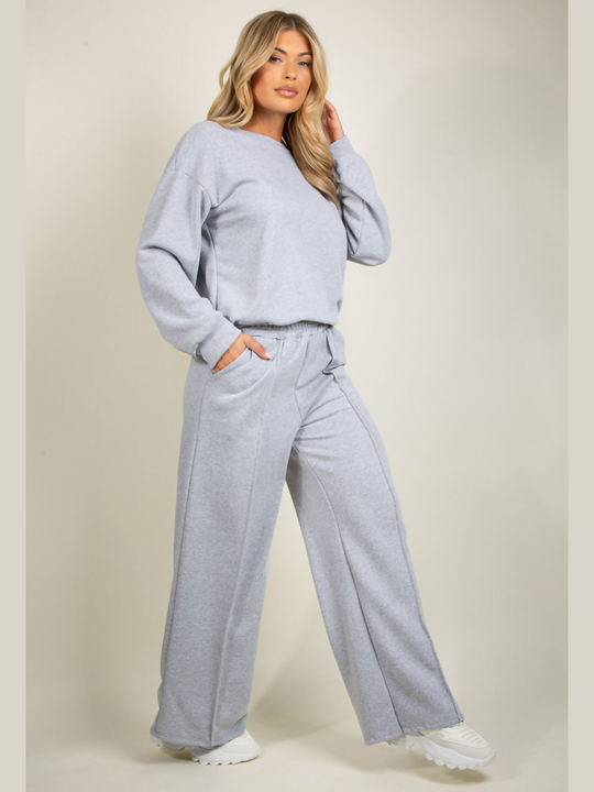 Full length shot. Model wears grey round neck long sleeve jumper and elasticated wide leg matching joggers. The joggers has a pockets anddecorative seam. Model has blonde hair and stands slightly to the side and has one hand in her pocket and by her head.