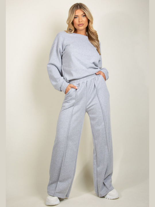 Full length shot. Model wears grey round neck long sleeve jumper and elasticated wide leg matching joggers. The joggers has a pockets anddecorative seam. Model has blonde hair, she faces the camera and places her hands in her pockets.