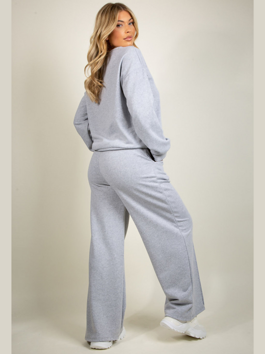 Full length shot. Model wears grey round neck long sleeve jumper and elasticated wide leg matching joggers. The joggers has a pockets anddecorative seam. Model has blonde hair, she stands with her back to the camera and has her hands in her pockets. The back of the joggers and the jumper can be seen.