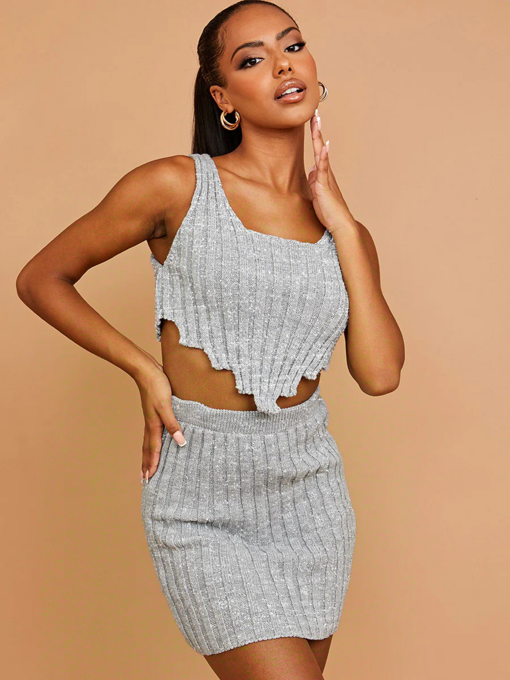 Model wears a grey knitted crop top with a jagged asymmetric hem and square  neckline. The top has a a silver flexedlurex detailing woven into the garment. The Top has an elasticated matching skirt. The model has brown skin and black hair that is pulled back into a long ponytail, she faces the camera, the front of the top and  the atching skirt can be seen. The model laces one hand on her hip and touches her chin with her other hand.