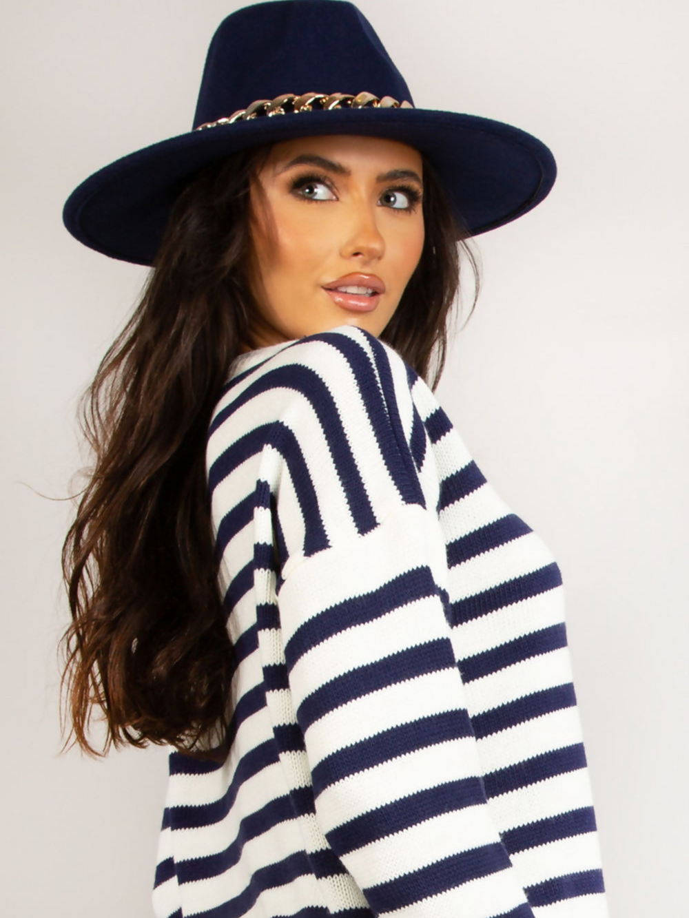 Model wears a luxe navy felt fedora hat with chunky gold chain design at the centre of the hat. Model stands to the side and looks off into the distance, the front of the hat can be seen.