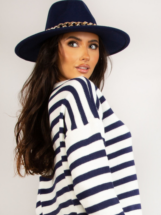 Model wears a luxe navy felt fedora hat with chunky gold chain design at the centre of the hat. Model stands to the side and looks off into the distance, the front of the hat can be seen.