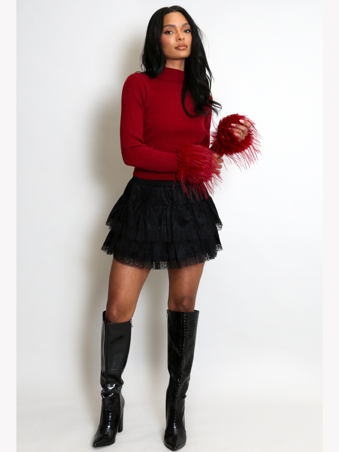 Full length shot. Model stands in front of a plain cream background. The model wears a red fine knit ribbed jumper with red faux fur cuffs and a black lace ruffled skirt. The model has light brown skin and long black hair, faces the camera, she wears black knee high boots and places her arms in front of her, the faux fur  cuffs can be seen.