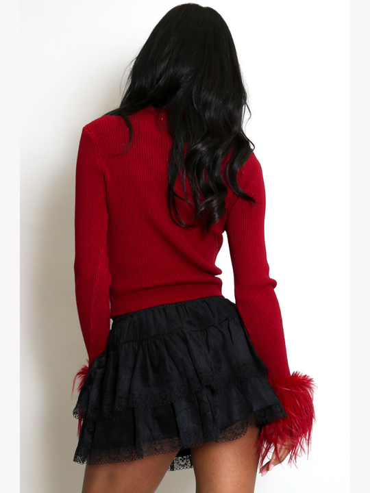 Model stands in front of a plain cream background. The model wears a red fine knit ribbed jumper with red faux fur  cuffs and a black lace ruffled skirt.  The model has light brown skin and long black hair, she stands with her back to the camera, the back of the fine knit ribbed jumper can be seen.