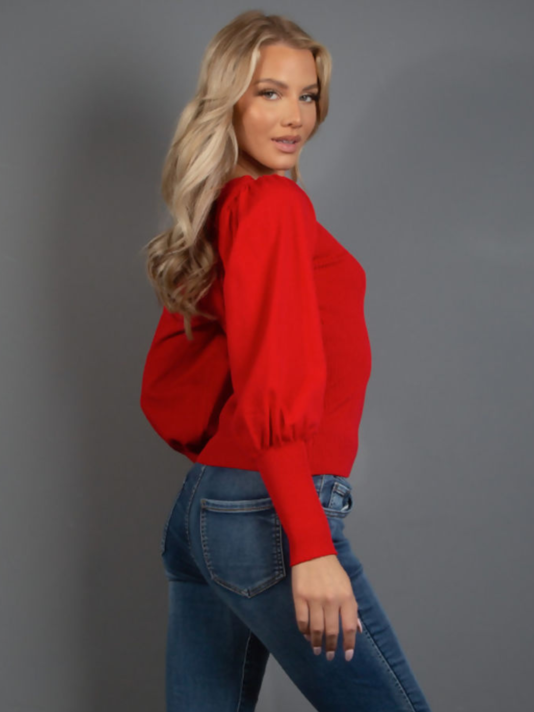 Female model wears a red long sleeve jumper with a rounded neck and gold military buttons at the front. The model has blonde hair and tanned skin,  she stands to the side and turns her head to look into the camera.