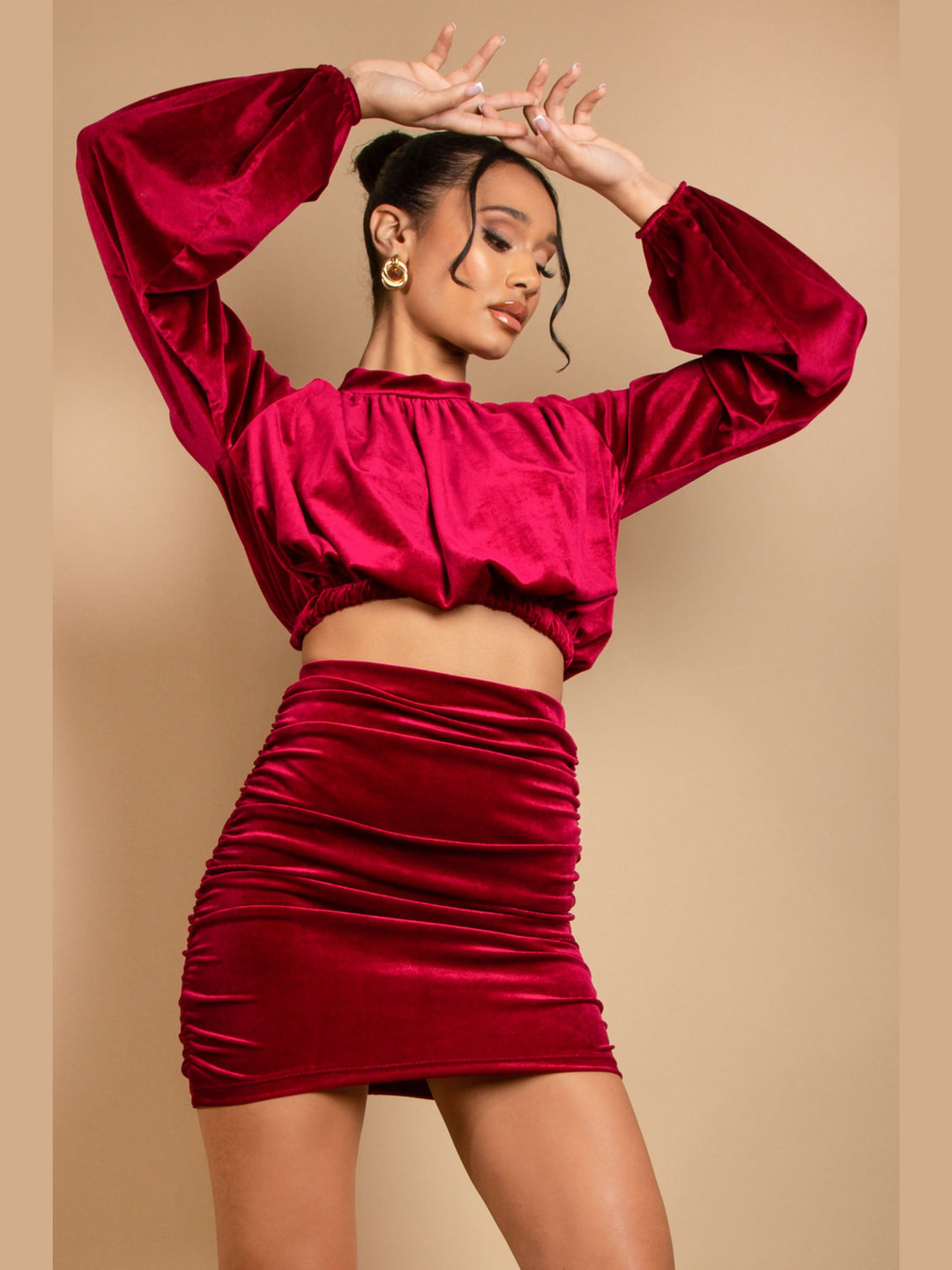 Model wears a long sleeeved red velvet crop top with a high neck. She also wears a matching  ruched mini skirt. The model faces forward and places her hands above her head and looks down.
