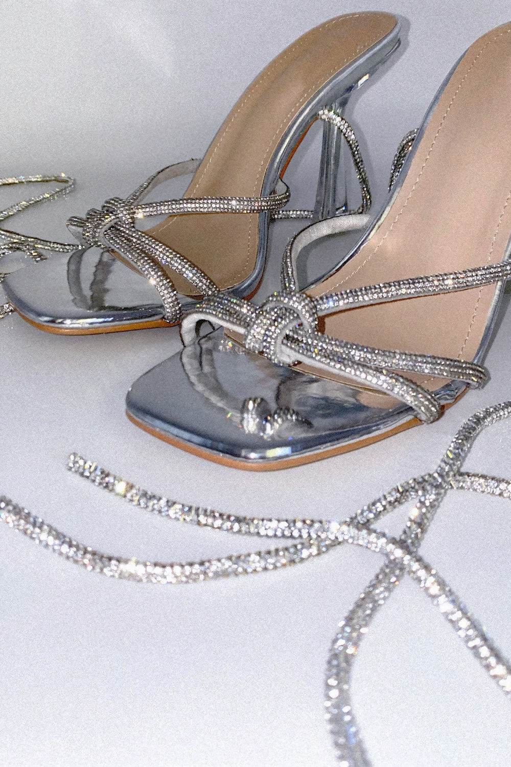 Close up of silver patentt leather look muled shoe, with open toes and silver diamante cross over straps. The shoe also has a silver diamante strap that  is designed to fasten along the shin