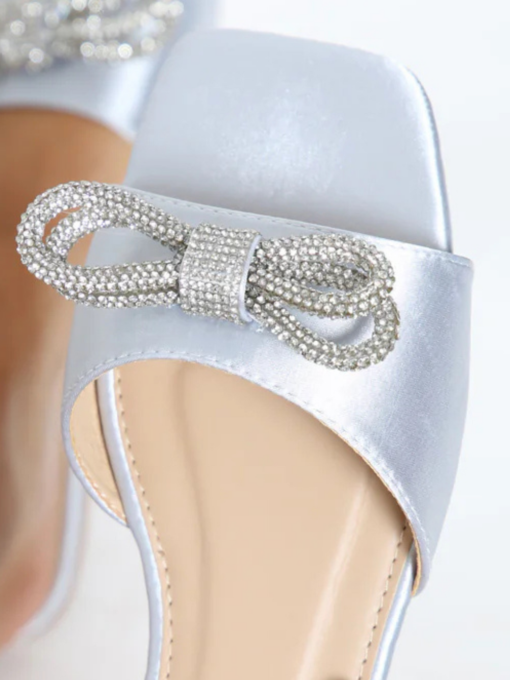 Closeup of diamante bow at the front of the flat shoe. Women wears a pair of silver flat satin shoes with diamante bow detailing. The design of the diamante bow is visible, demonstrating the central dimante ring at its centre.