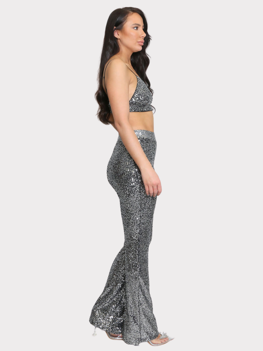 Full length shot. Model wears silver sequin bralette with a v neck neck line and adjustable straps. The sequined matching trousers have an elasticated waist. Model stands to the side. The whole of the co-ordinated set can be seen. Model wears sparkly diamante shoes.