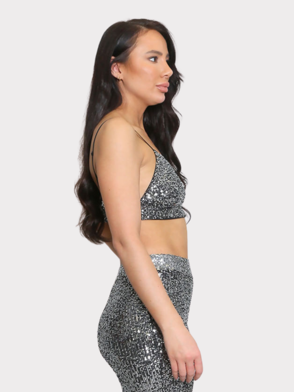 Model wears silver sequin bralette with a v neck neck line and adjustable straps. The sequined matching trousers have an elasticated waist.  Model stands to the side. The bralette and adjustable straps can be seen. Model stands with her hands by her sides and her lon brown wavy hair can be seen.