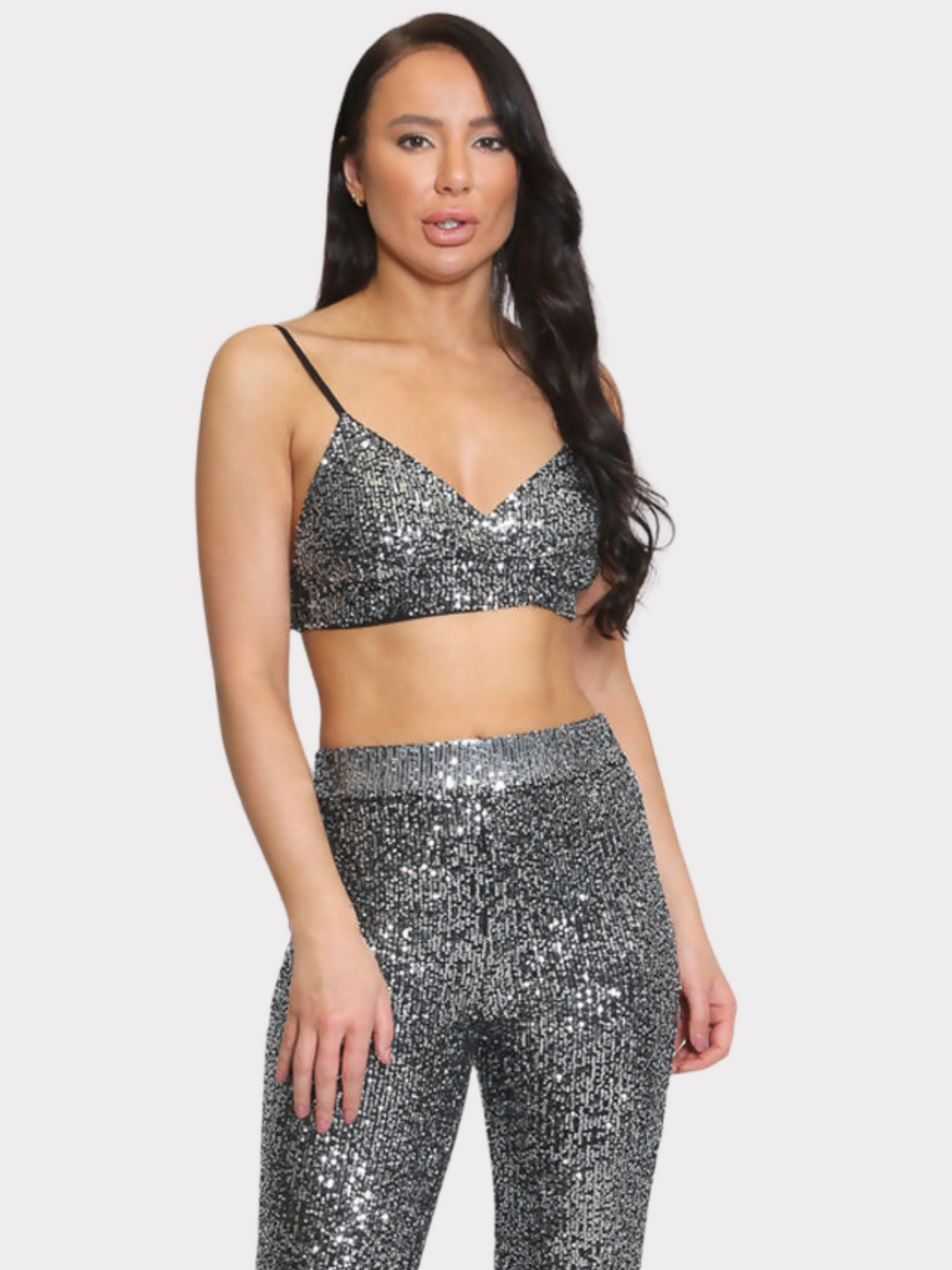 Model wears silver sequin bralette with a v neck neck line and adjustable straps. The sequined matching trousers have an elasticated waist. Model faces forward and places one hand on her thigh and the other by her side.