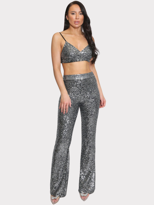 Full length shot. Model wears silver sequin bralette with a v neck neck line and adjustable straps. The sequined matching trousers have an elasticated waist.  Model faces the camera and has one hand on her thigh and the other on her hip. She wears a sparkly shoes.