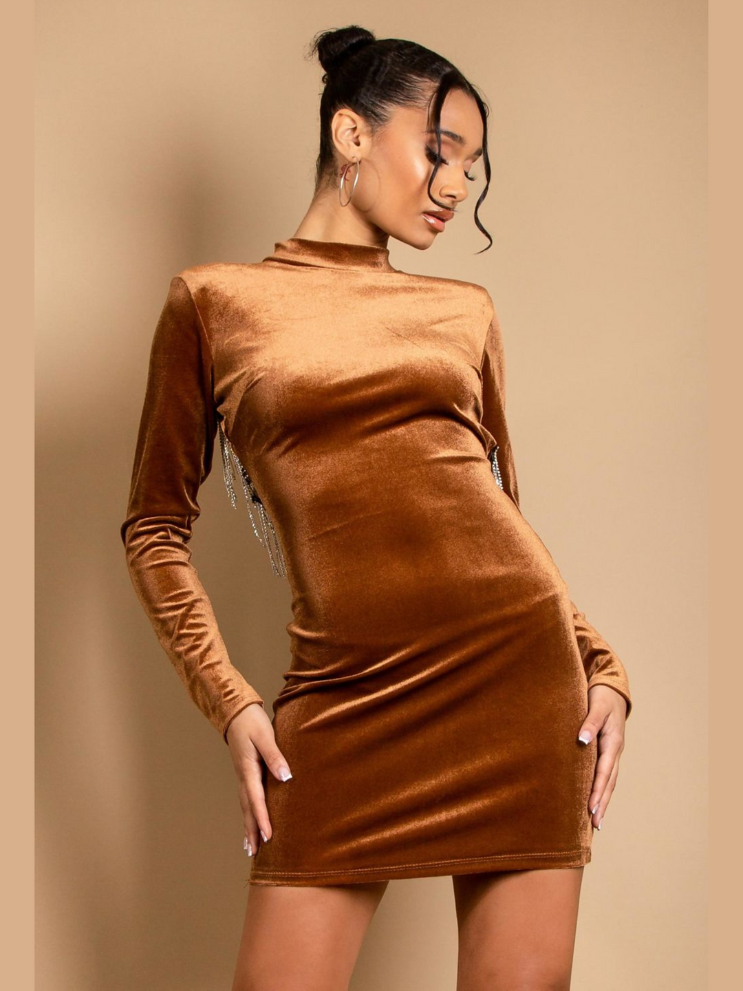 Model wears a velvet mini dress in a tanned colour. The mini dress has a bodycon fit and velvet elastic, with a mock neck, long sleeves and a cut out back with diamante trim. Model faces forward with her hands by her sides. The diamante tassles situated at the back can ne seen hanging down. Model looks down.