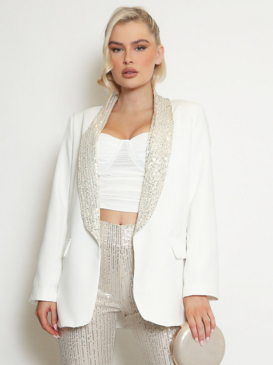 Model wears an oversized blazer with sequin lapel  and single front button fastening. Model faces the camera and holds a clutch bag.