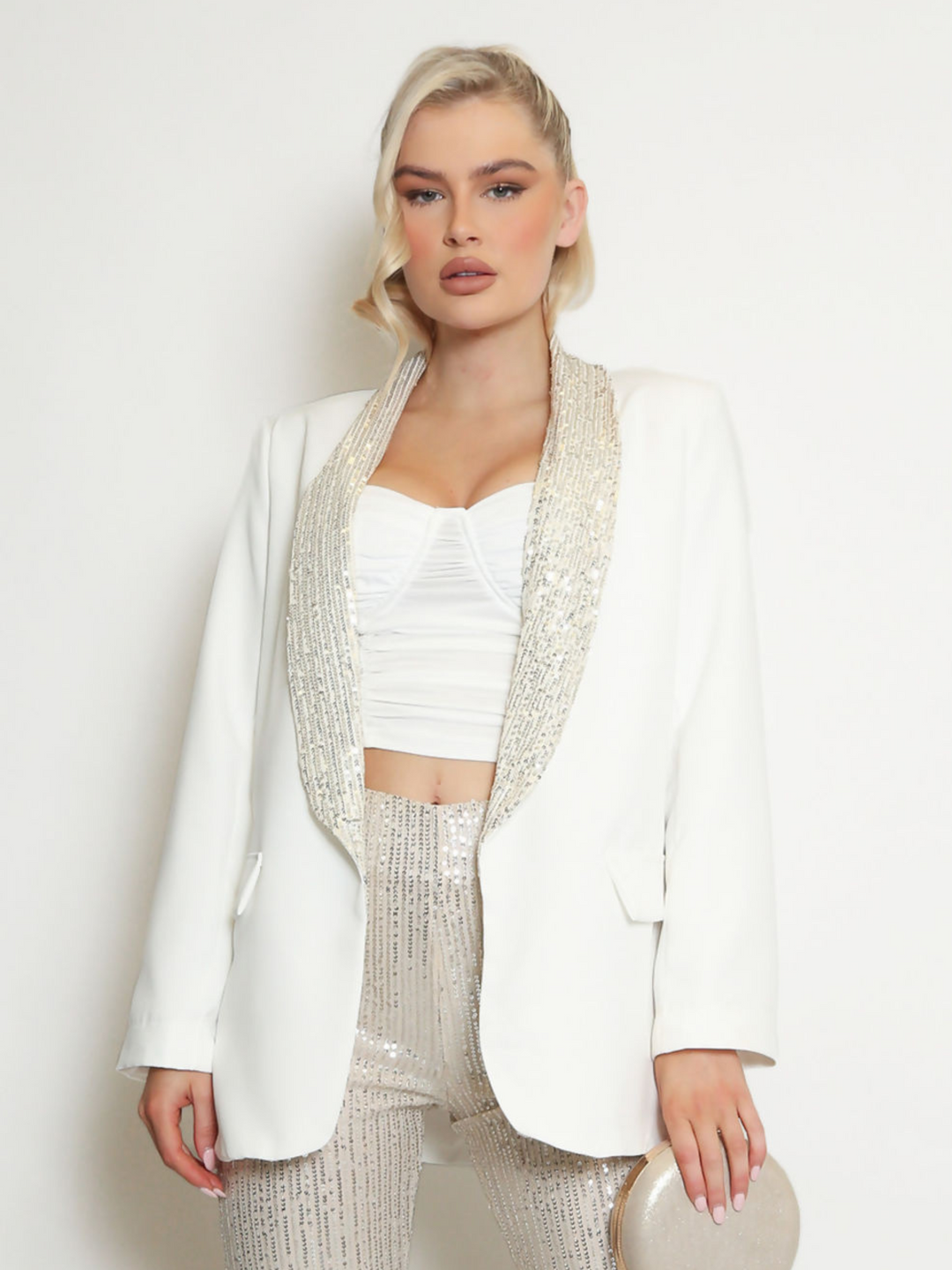 Model wears a white oversized blazer with sequin lapel and single front button fastening. Model faces forward and holds a silver clutch bag.