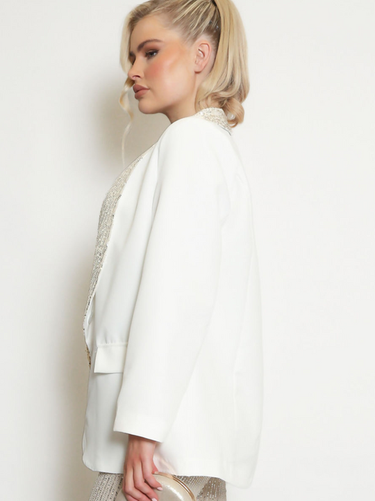 Model wears a white oversized blazer with sequin lapel and single front button fastening. Model syands to the side. The side of the white oversized blazer can be seen. The silver  sequin lapel trim is visible.