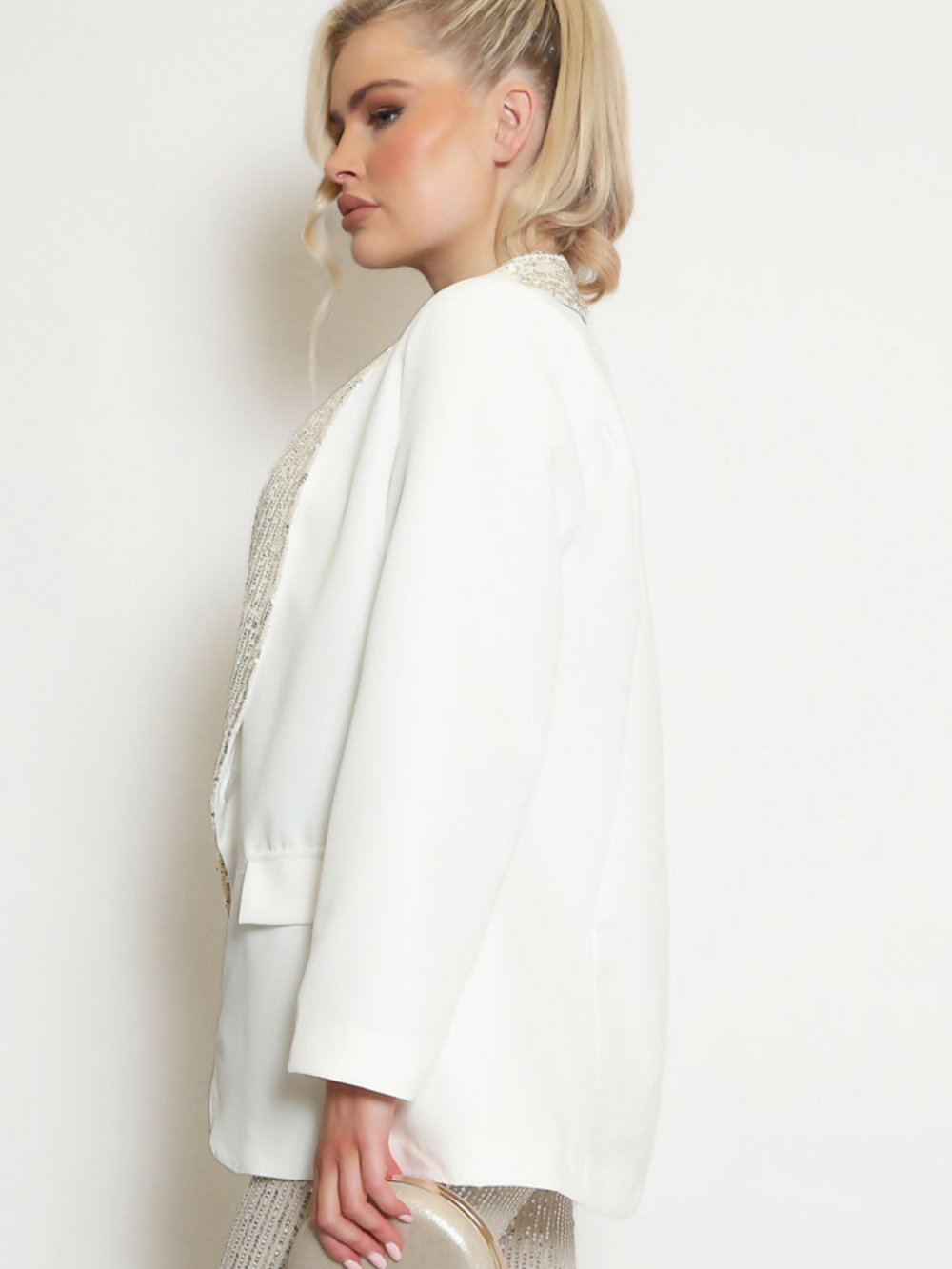 Model wears a white oversized blazer with sequin lapel  and single front button fastening.  Model stands to the side holding a clutch.