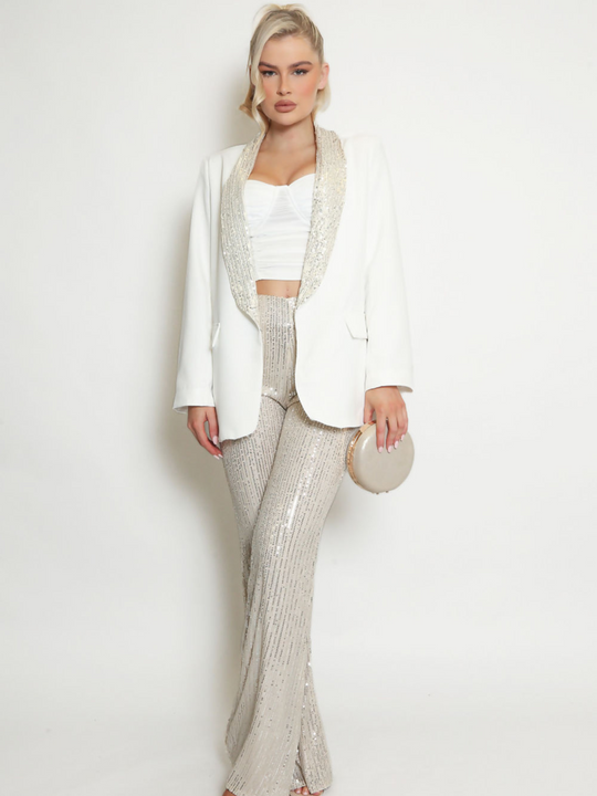 Model wears a white oversized blazer with sequin lapel  and single front button fastening.  Model faces forward,  she is wearing a white crop top and sequin trousers and holds a silver clutch purse.