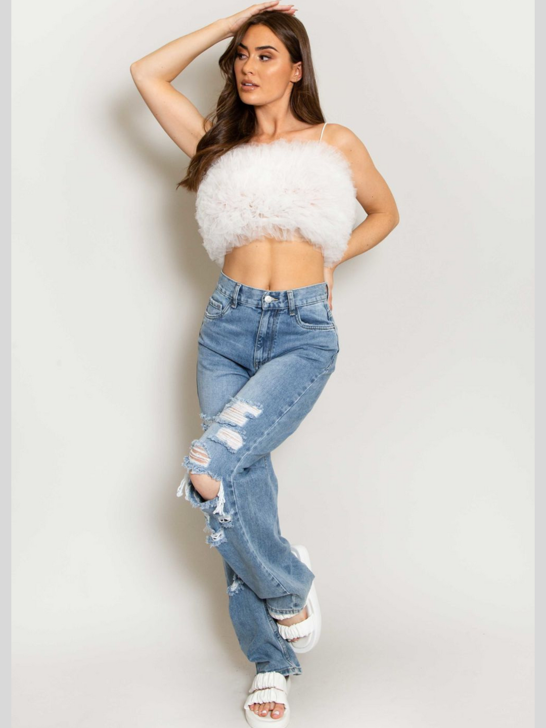 Full length shot. Model wears  a white organza ruffled crop top ,with spaghetti strap and zip back fastening. Model has tanned skin and long brown hair.  Model wears a denim jean, she llifts one leg slightly raised and places one hand on her head.