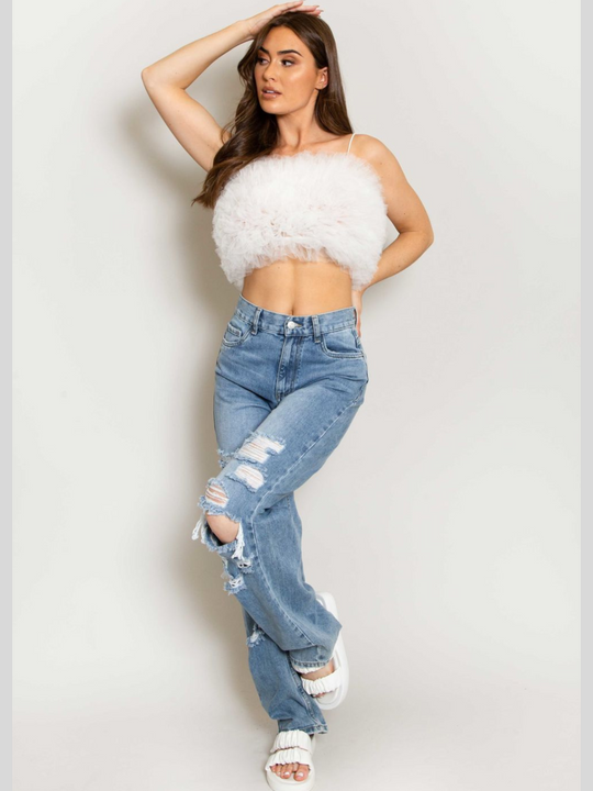 Full length shot. Model wears  a white organza ruffled crop top ,with spaghetti strap and zip back fastening. Model has tanned skin and long brown hair.  Model wears a denim jean, she llifts one leg slightly raised and places one hand on her head.