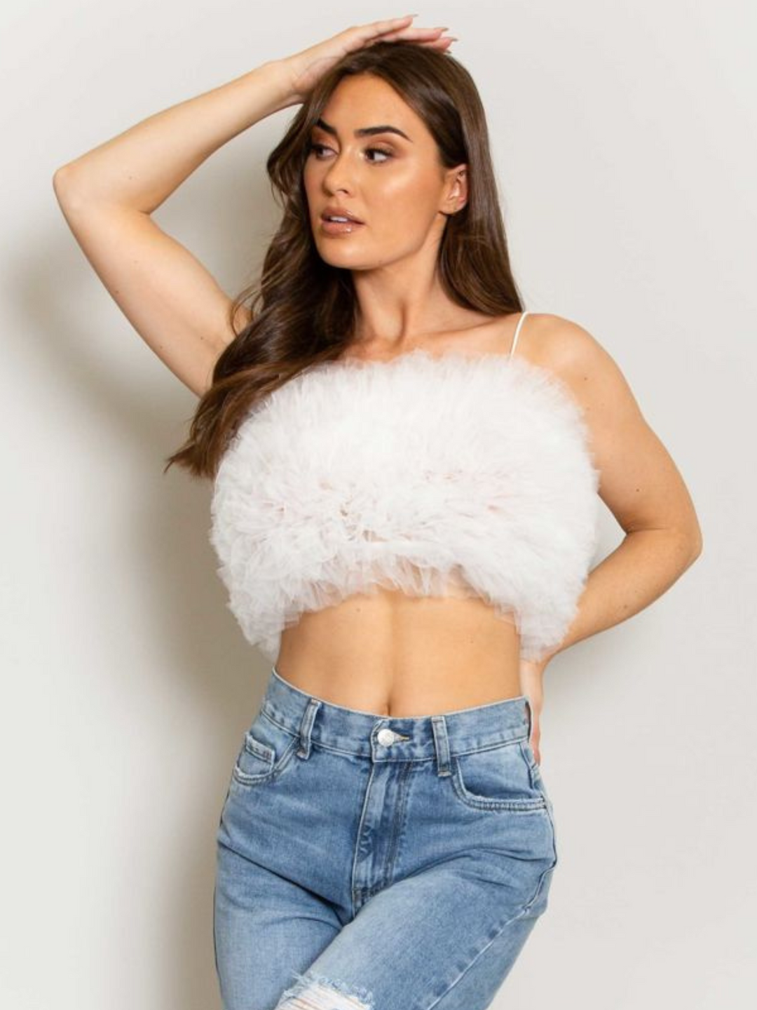 Model wears  a white organza ruffled crop top ,with spaghetti strap and zip back fastening. Model has tanned skin and long brown hair. She looks to the side and places one hand on her hip and the other behind her head.