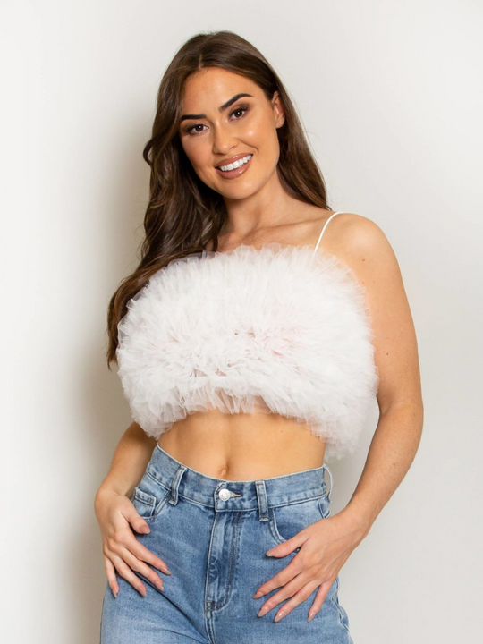 Model wears  a white organza ruffled crop top ,with spaghetti strap and zip back fastening. Model has tanned skin and long brown hair. Model faces the camera an places both hands in front of her and tilts her head sllightly to the side and smiles.