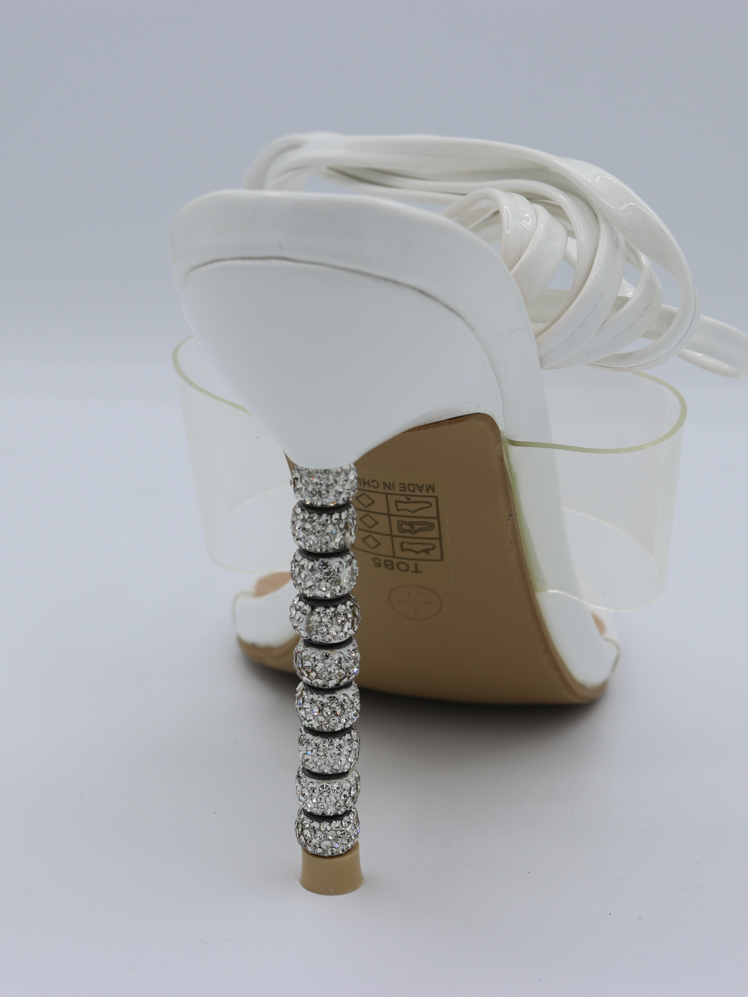 White strappy mules shoes with diamante stiletto heel. This is a statement shoe with a diamante heel. The image shows the back of the shoe and the white straps can be seen piled on top of the heel.