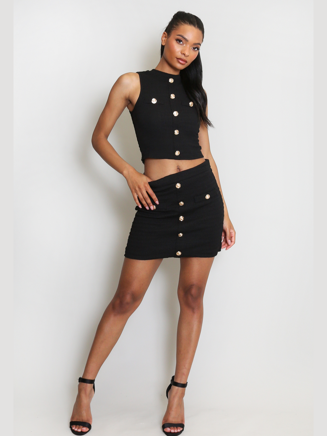 Full length shot. Model wears  luxe black plisse mini skirt with gold military button detailing to the front of the dress. The model has brown skin and long black hair, she stands  facing forwardwith one leg to the side the side and places one hand by her side and the other at the skirt's waist.