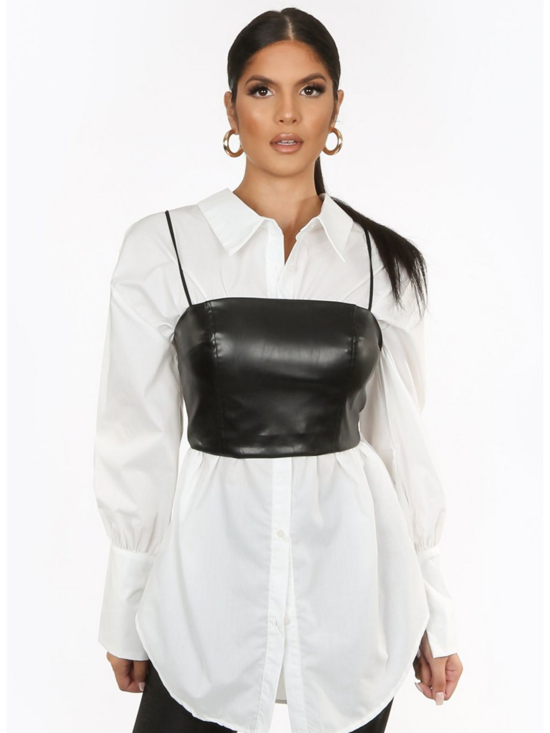 Model wears a pu leather look crop top cami with spaghetti straps and zip back fastening. The model wears a shirt underneath the crop  top. The model stands with her hands by her side.