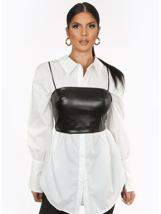 Model wears a pu leather look crop top cami with spaghetti straps and zip back fastening. The model wears a shirt underneath the crop  top. The model stands with her hands by her side.