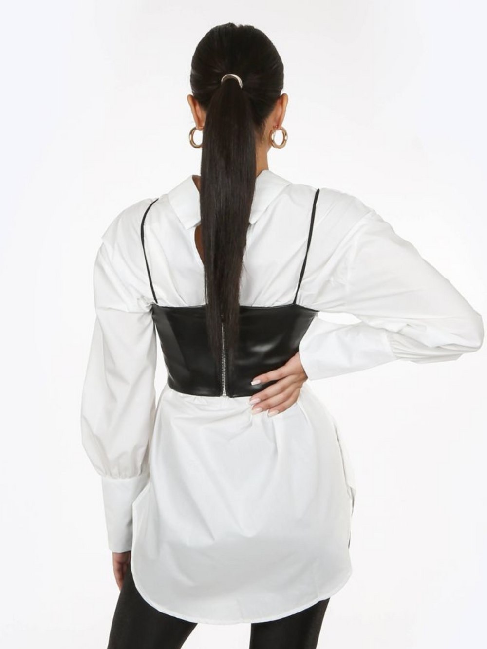 Model wears a pu leather look crop top cami with spaghetti straps and zip back fastening. The model wears a shirt underneath the crop  top. The model stands with her back to the camera, the back of top can be seen, including their zip back fastening.