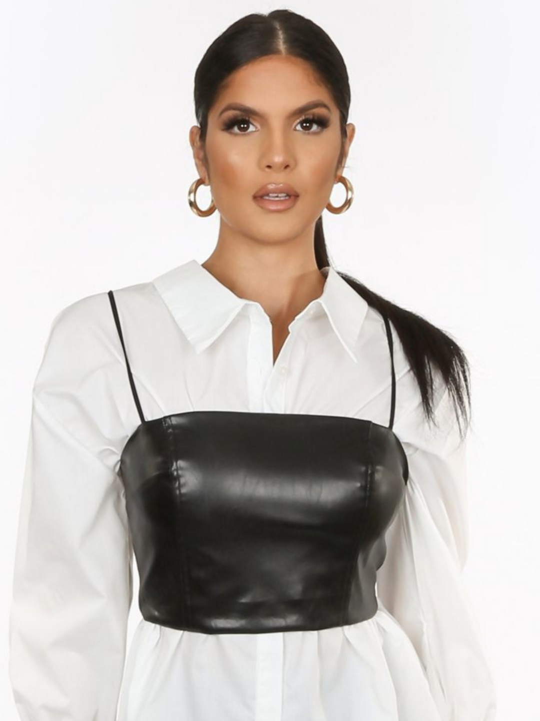 Model wears a pu leather look crop top cami with spaghetti straps and zip back fastening. The model wears a shirt underneath the crop  top. The model stands with her hands by her side. Model wears gold hoop earings.