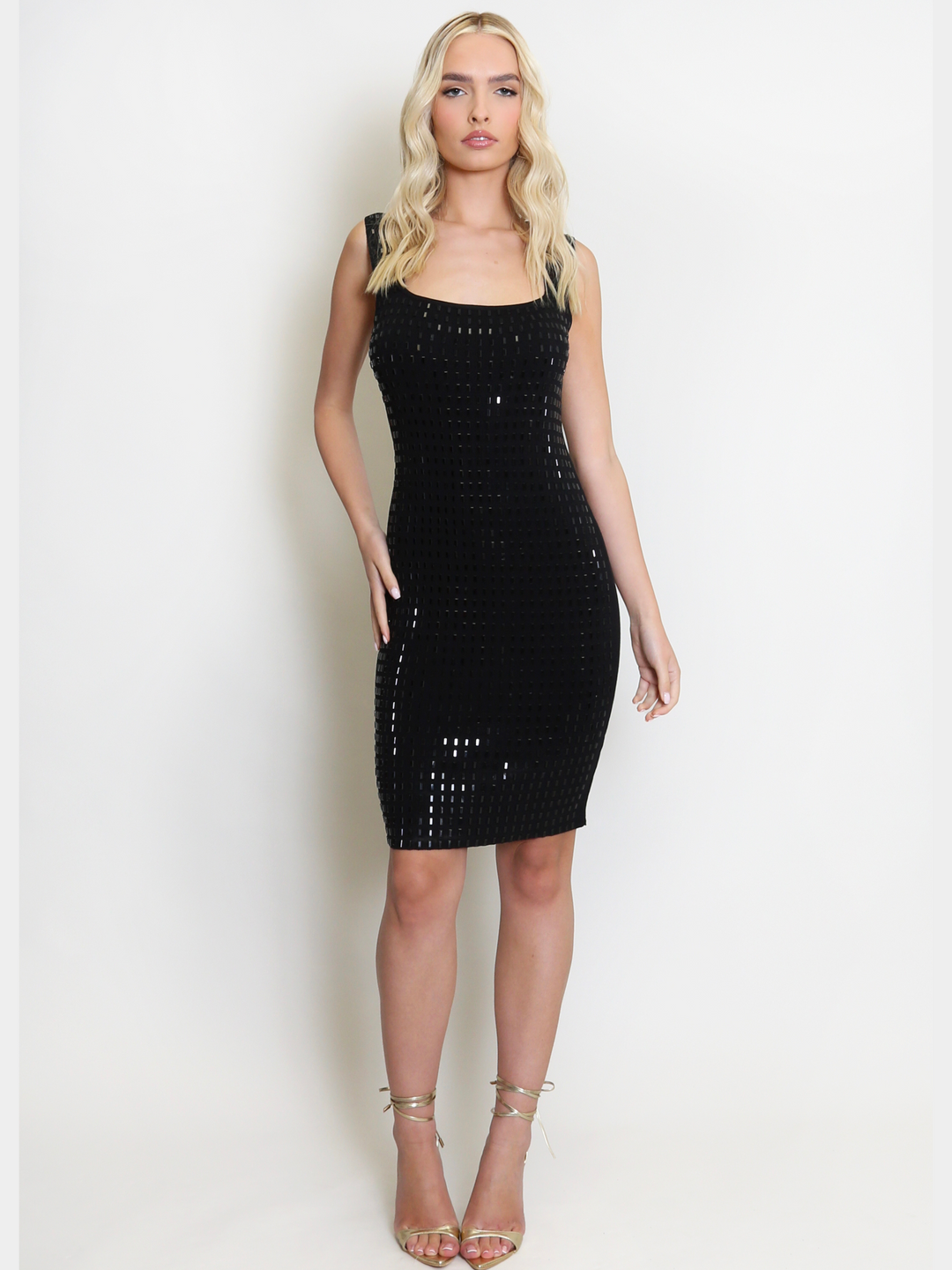 Full length shot. Female model wears a black midi dress and stands in front of a plain background. The black midi dress has a  silver rhinestone embelished design to the front and a square cut neckline. The model has tanned skin and long blonde hair, she faces forward, placing one hand on her hip and the other by her side, she wears gold lace up heels on her feet.