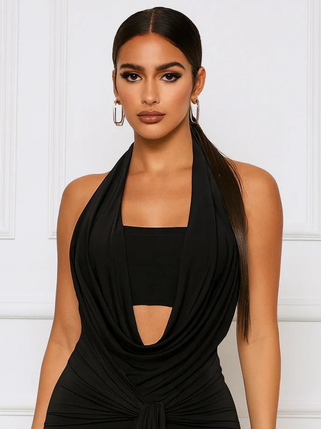 Mid length shot. Female model stands before a panelled white wall. The model wears slinky black mini dress with a plunging cowl front and a  rape tie front.