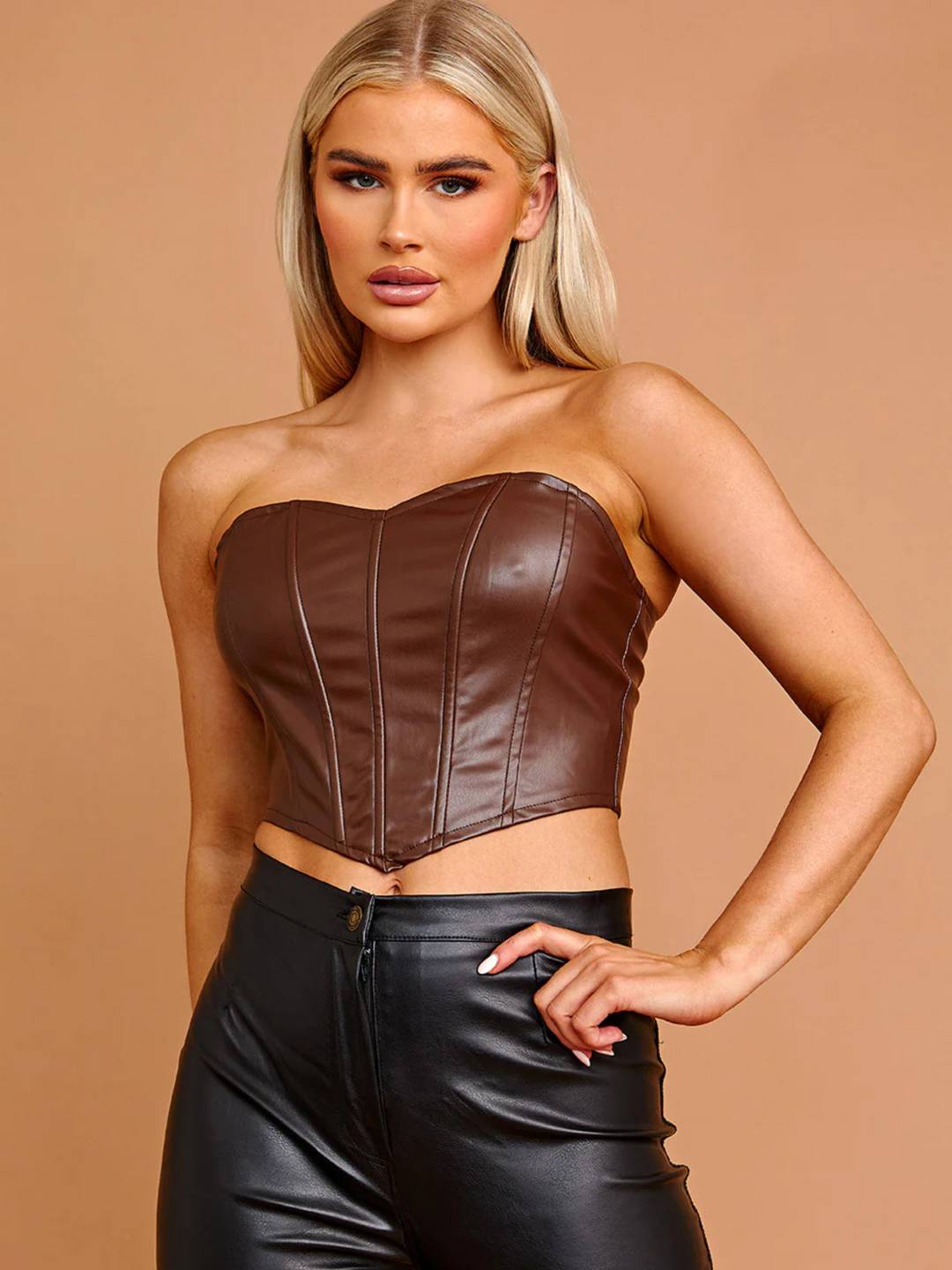 Model wears a brown pu leather bandeau crop top, that has a sweet heart neckline and corset style panellig to the front and zip back fastening to the rear. The model has tanned skin and long blonde hair, she faces forward and places one hand on her hip and the other behind her back.