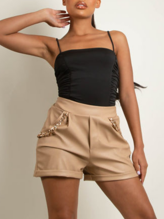Mid length shot. Model wears beige pu leather shorts with front side pockets and gold chain drape detailing at either side of her shorts.