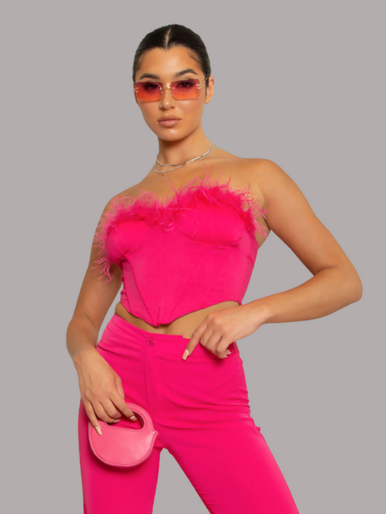 Model wears bright fuschia pink crop top, with fuschia pink feathers and zip bak fastening. The model has tanned skin, with black hair that is slicked back. The model faces forward, placing one hand on her waist and holds a pink mini bag in her other hand.