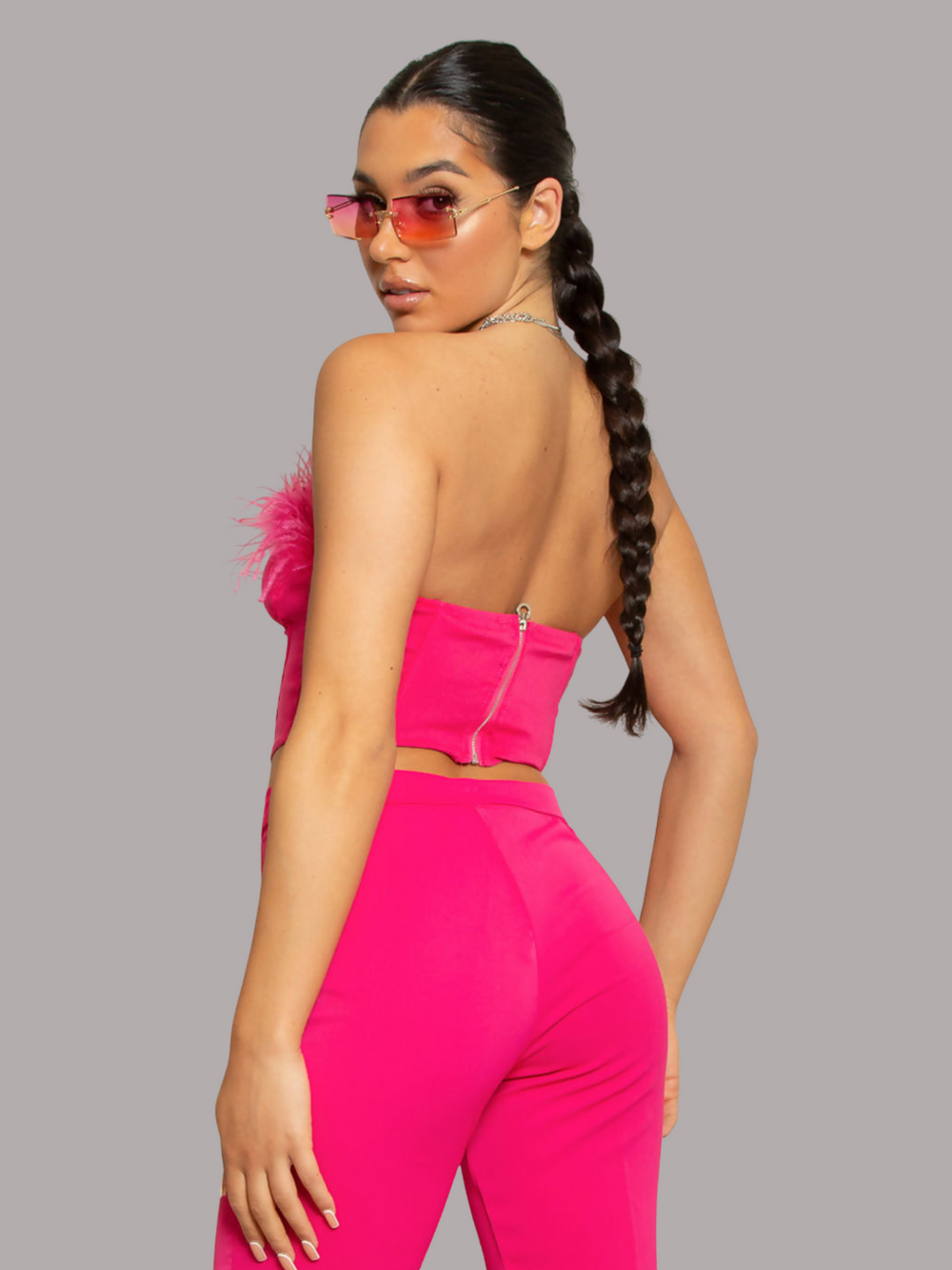 Model wears bright fuschia pink crop top, with fuschia pink feathers and zip bak fastening. The model has tanned skin, with black hair that is slicked back plaited pony tail. The model stands with her back to the camera, the back of the top can be seen.